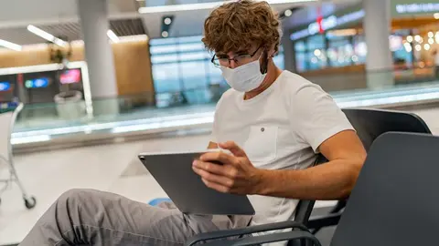 Man in respirator mask is waiting next plane at the airport and using  tablet. Coronavirus COVID-19 concept.
